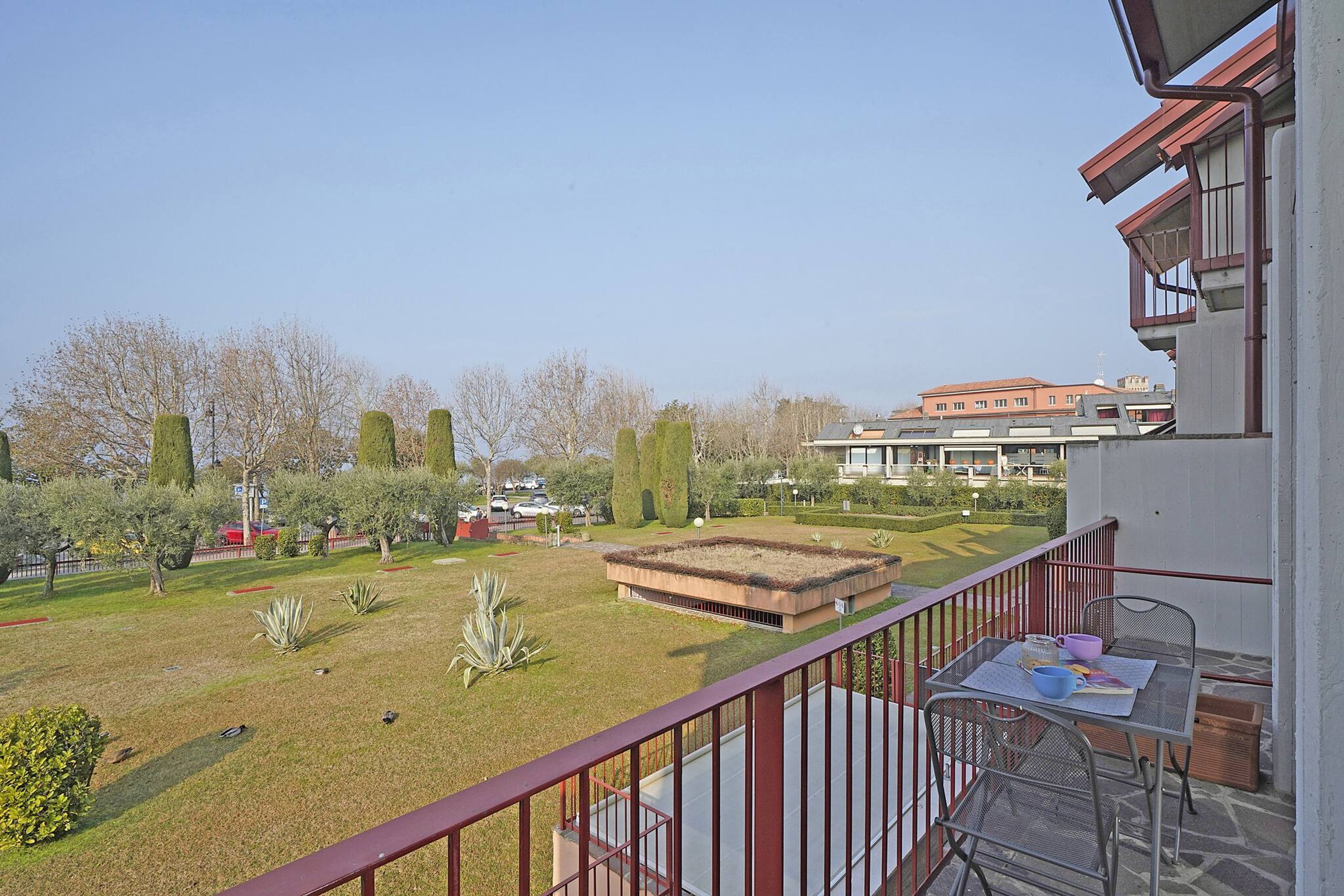 A balcony with a table and chairs, overlooking a well-maintained garden with various plants and a building in the background.
