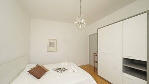 Ein Schlafzimmer mit einem Bett, einem braunen Kopfkissen, einer weißen Daunendecke, einer Kandelaberleuchte, einem Kleiderschrank und einem gerahmten Bild an der Wand.