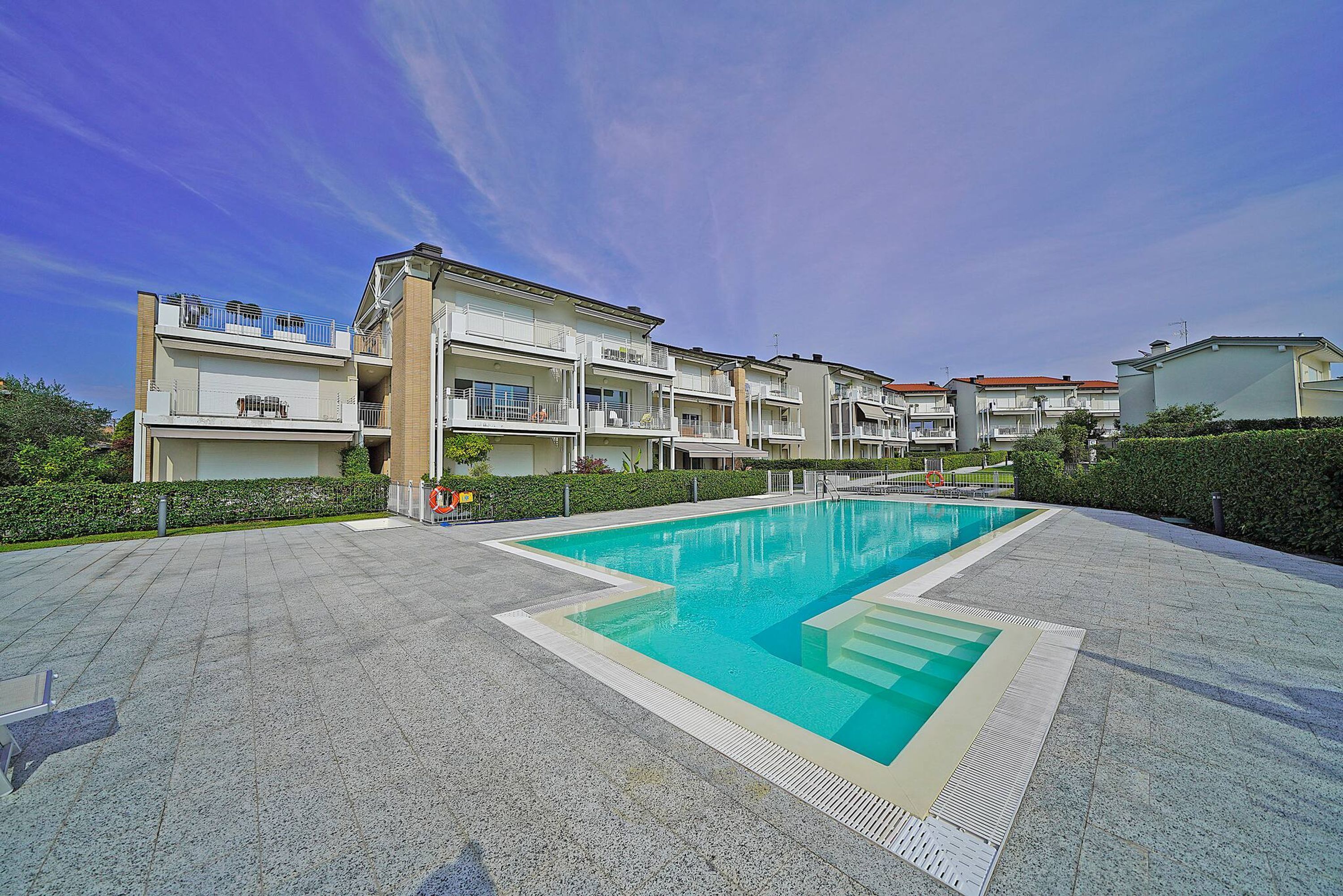 Un espace piscine avec une piscine à l’eau d’un bleu limpide, entourée d’une allée pavée, et jouxtant un immeuble résidentiel à plusieurs étages doté de balcons.