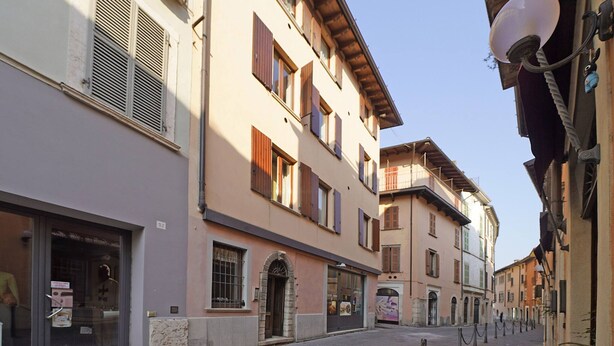 A narrow cobblestone street lined with multi-story buildings, featuring ground-floor shops and upper-floor residential or commercial spaces. - Casa Annalisa in Sal (Salò)