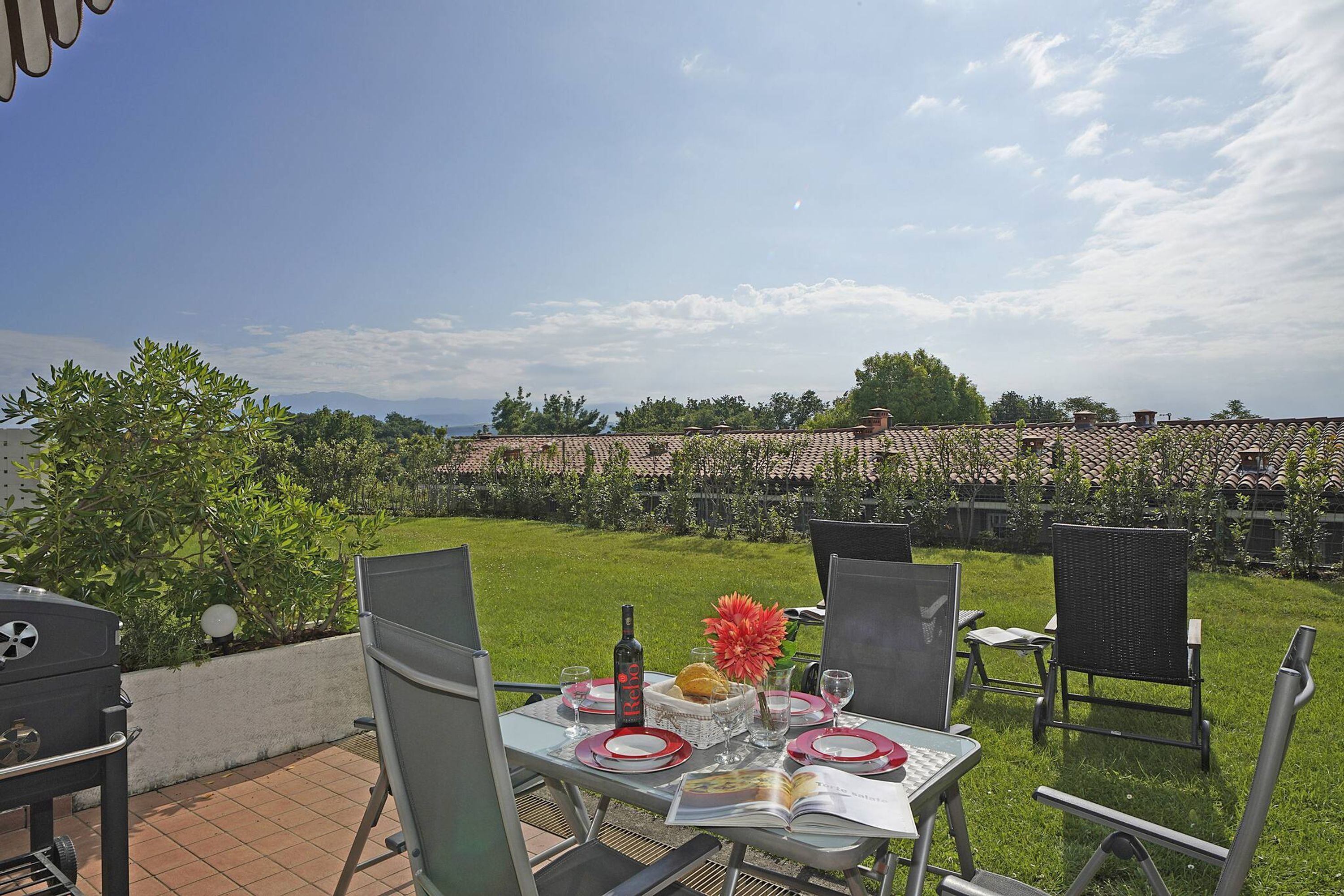 An outdoor patio with a table set for dining, featuring a bottle, glasses, and a book.