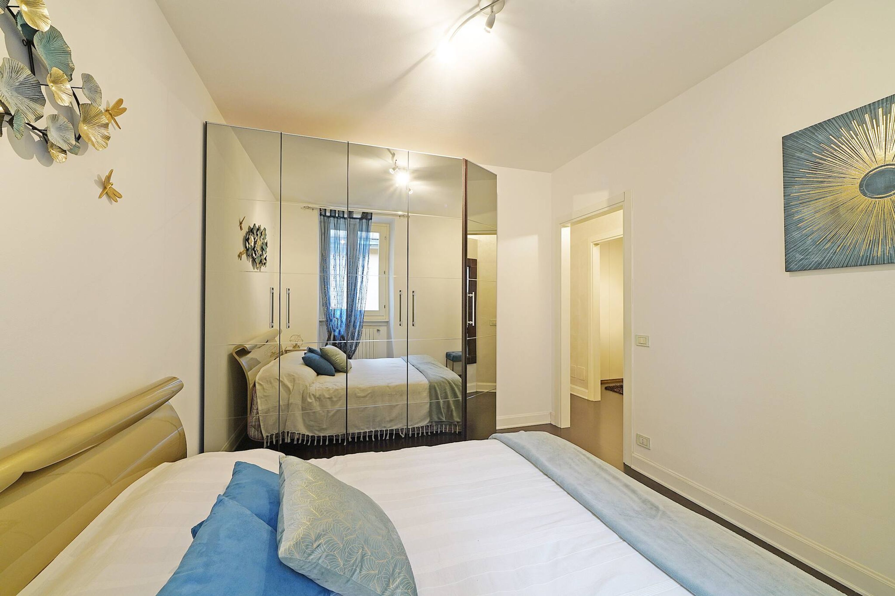 Une chambre à coucher comprenant un lit, un grand miroir, une œuvre d’art murale et une porte donnant sur une autre pièce.