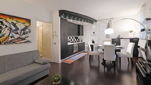 A modern living room with a grey sofa, a dining area with white chairs, a kitchen with dark cabinets, and a large painting on the wall. - Downtown Apartment in Sal (Salò)