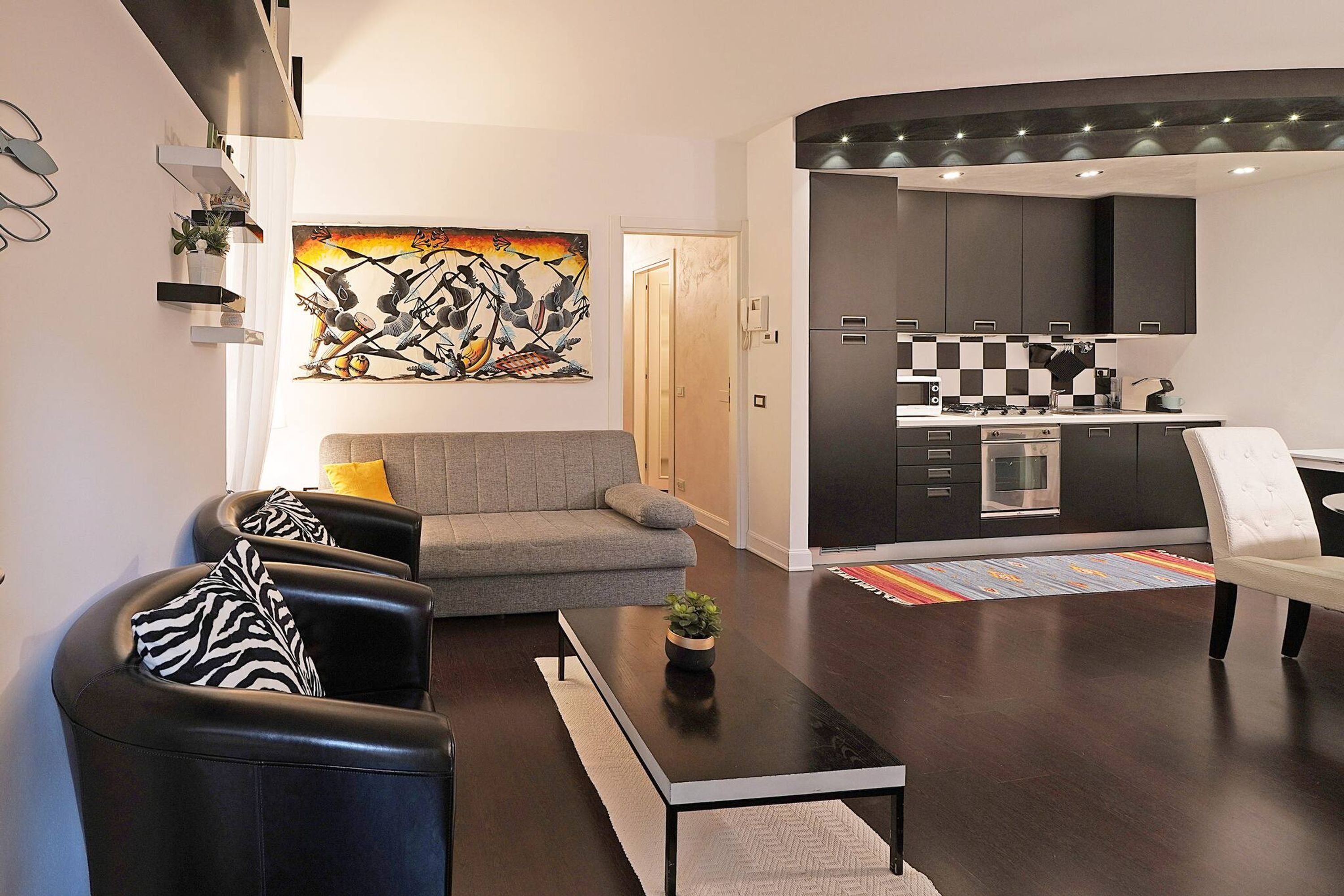 A modern living room with a grey sofa, black leather armchairs, and a coffee table. A kitchen with dark cabinets and a checkered backsplash is visible through an open door.