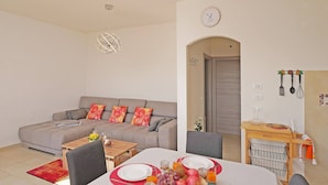 Un salon moderne avec un canapé gris, une table à manger dressée avec des assiettes, des verres et des fruits. Il y a une horloge au mur et un coin cuisine avec des ustensiles suspendus au-dessus.
