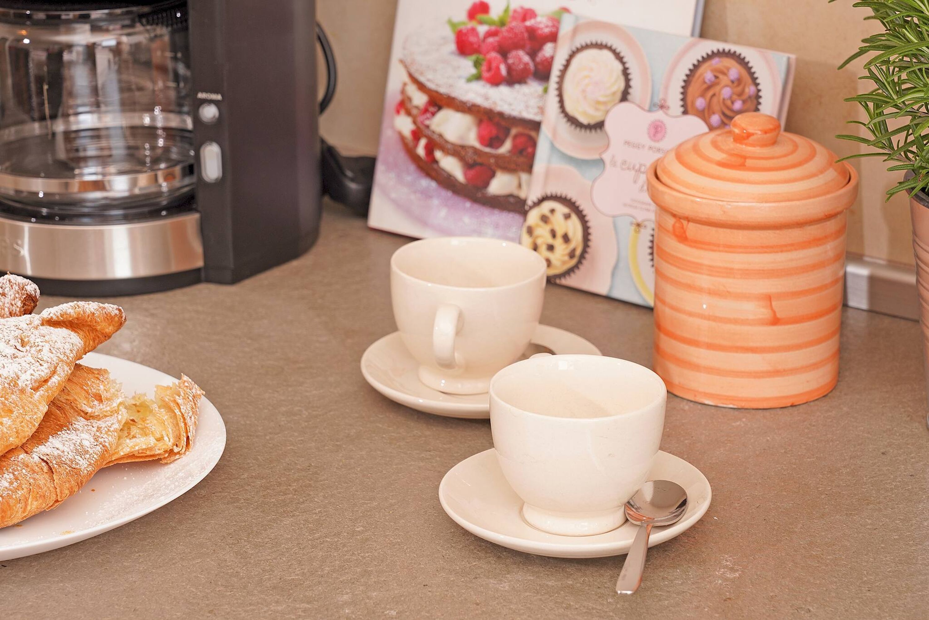 Une table préparée pour le petit-déjeuner avec des viennoiseries, deux tasses blanches avec leurs soucoupes, une cuillère, une cafetière et un pot décoratif.