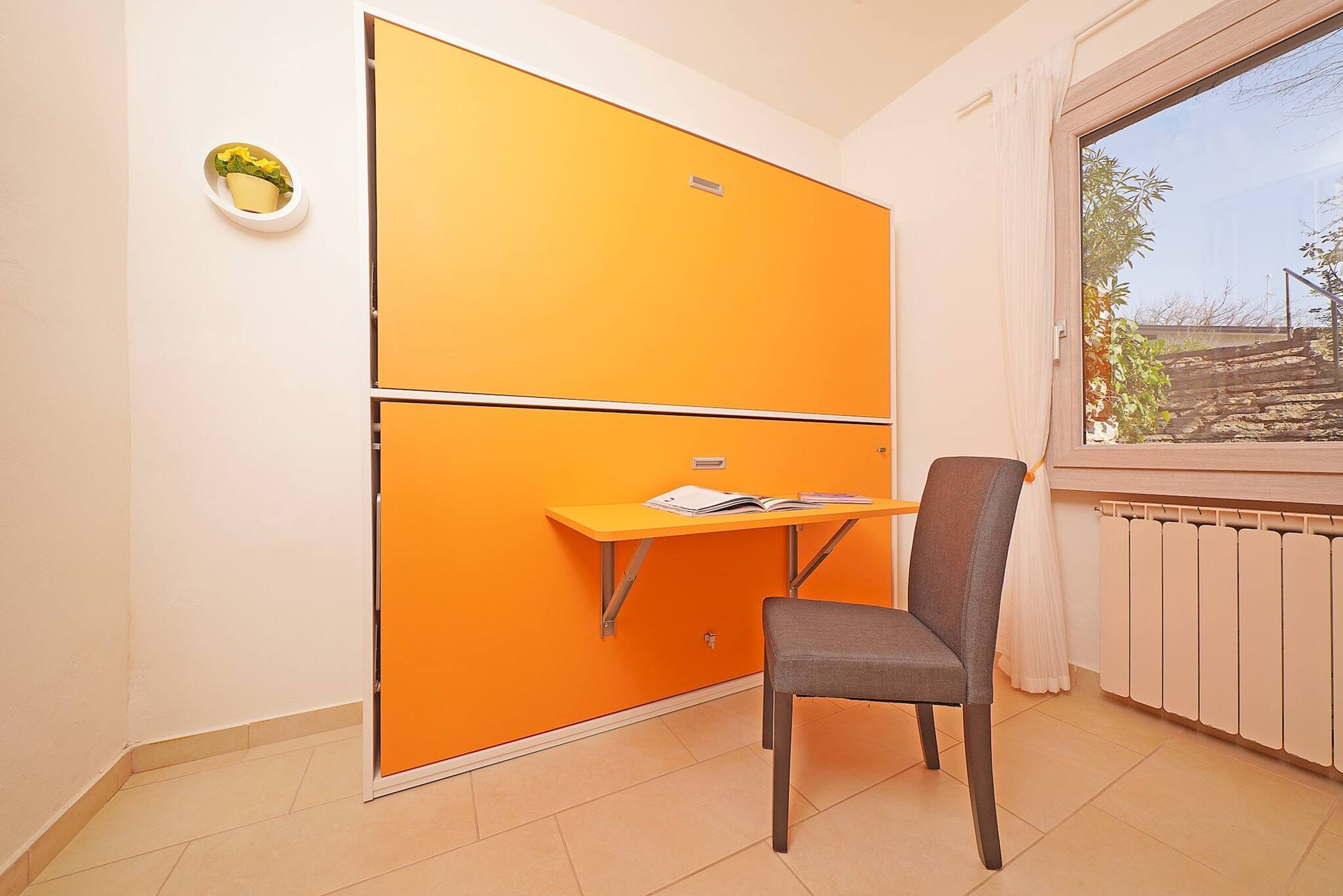 Une pièce avec une grande armoire orange, une petite table avec une chaise et une fenêtre donnant sur un immeuble et des arbres.