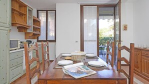 A dining area with a wooden table set for four, surrounded by wooden chairs. A wicker basket and plates are on the table, and a magazine is open. The room has white walls, a wooden cabinet, and a window with a view of the outside. - Villaggio dei Fiori - Fiordaliso (Sirmione)