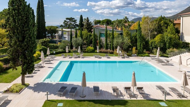 Piscina rectangular con sillas de descanso y sombrillas, rodeada de vegetación y edificios.