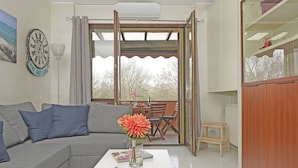 A living room with a grey sofa, a coffee table with magazines, a vase with flowers, a clock, and a view of the outside balcony. - Casa Rudi in Sirmione (Sirmione)