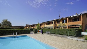 A swimming pool with a wooden deck, surrounded by a row of yellow buildings with balconies and a hedge. - Casa Rudi in Sirmione (Sirmione)