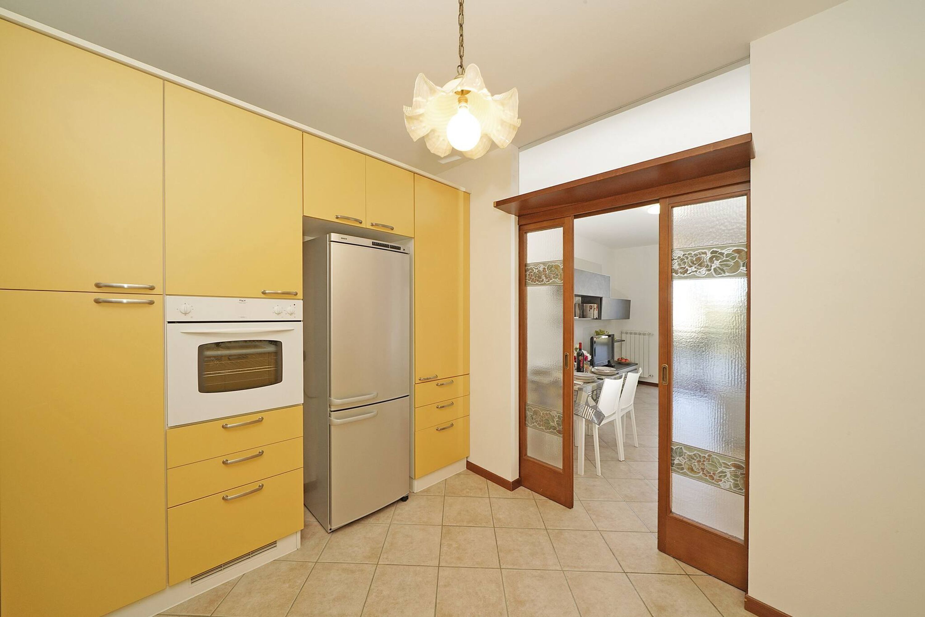 Una cocina con armarios amarillos, un refrigerador de acero inoxidable y un horno. Hay una puerta de madera con un panel de vidrio que da a otra habitación.