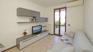 Ein modernes Wohnzimmer mit grauem Sofa, einem Flachbildfernseher auf einer eingebauten Konsole, einem kleinen Tisch mit Blumen in einer Vase und einer Schiebetür aus Glas, die zu einem Außenbereich führt.