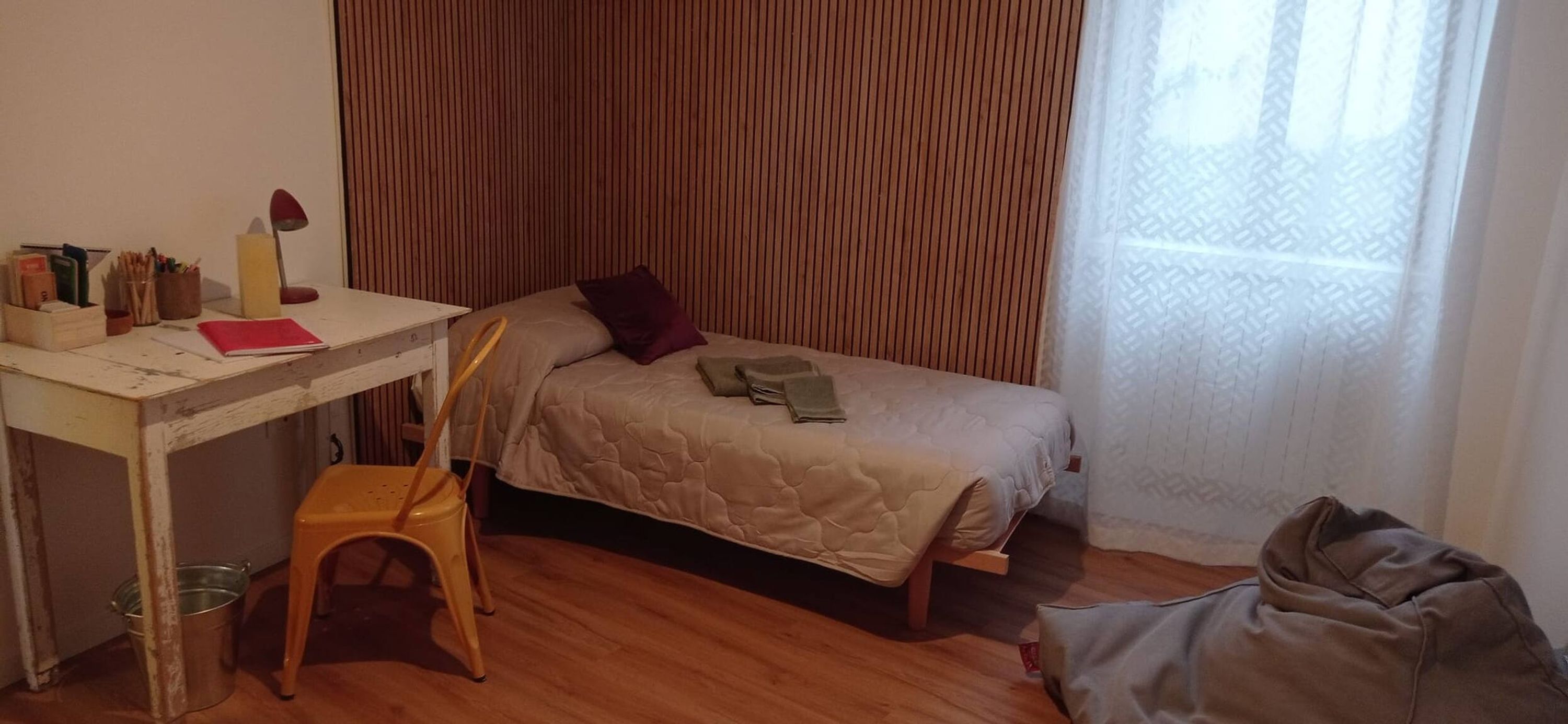 A small, tidy room with a wooden desk, a chair, a bed with a quilted cover, and a window with sheer curtains.
