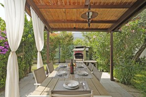 Eine hölzerne Pergola im Freien mit einem Esstisch für vier Personen, umgeben von üppiger Bepflanzung und einem Backofen im Hintergrund.