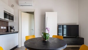 A modern kitchen with a built-in oven and coffee machine, a dining area with a round table and yellow chairs, and a flat-screen TV mounted on the wall. - Fasanella 50 in Toscolano-maderno (Toscolano Maderno)