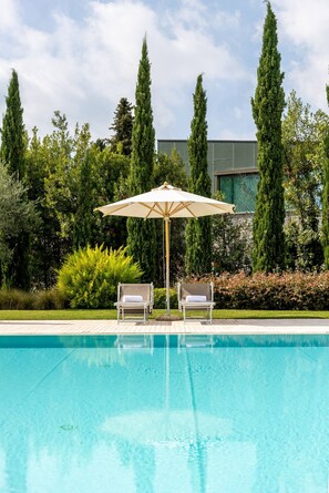 A swimming pool with two lounge chairs and a canopy, surrounded by tall trees and a modern building. - Fasanella 50 in Toscolano-maderno (Toscolano Maderno)