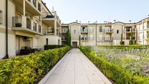 A pathway flanked by neatly trimmed hedges and lavender bushes, leading to a row of beige apartment buildings with balconies and air conditioning units. - Fasanella 50 in Toscolano-maderno (Toscolano Maderno)