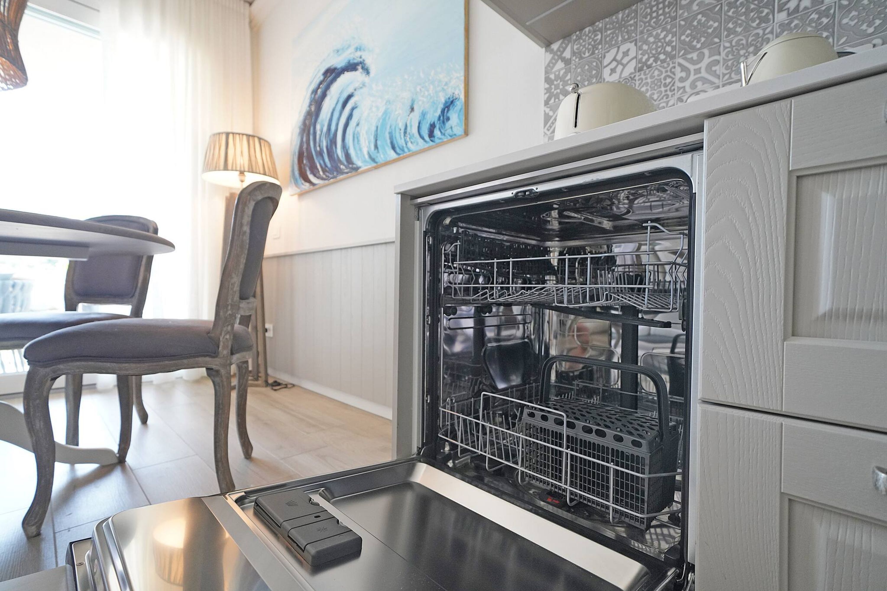 An open dishwasher with various kitchenware inside, a dining table with chairs, a lamp, and a painting on the wall.