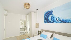 Una habitación de hotel moderna con una cama grande, un cuadro de una ola, una mesita de noche con una lámpara y un armario.