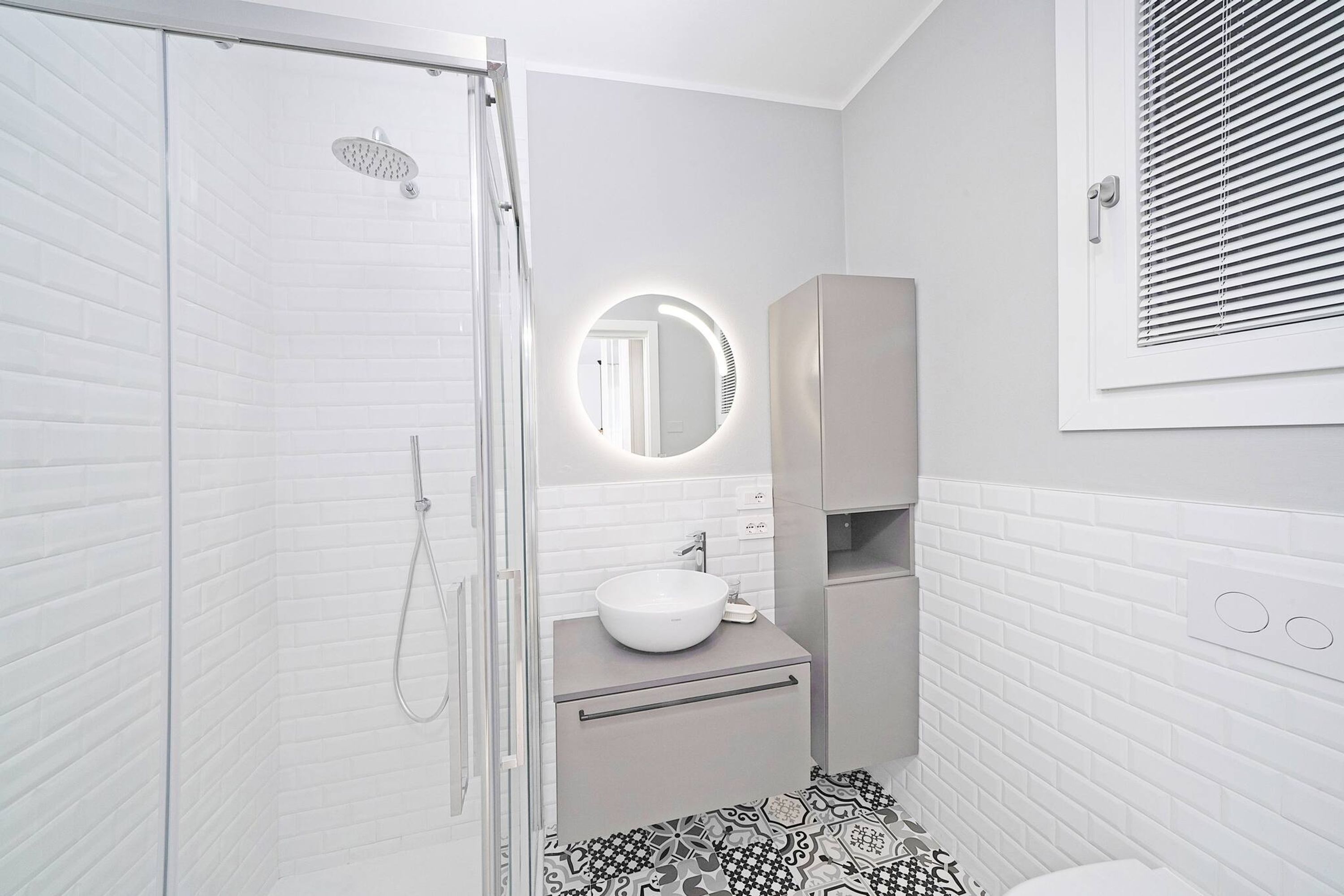 Une salle de bain moderne avec une douche à parois de verre, un grand miroir rond doté d’un éclairage intégré, un lavabo blanc et des toilettes.