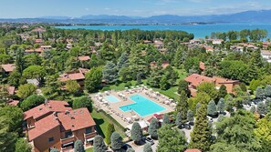 An aerial view of a residential area with a large swimming pool surrounded by lounge chairs and umbrellas, set against a backdrop of a lake and mountains. - Holiday Punta Gro in Sirmione (Sirmione)
