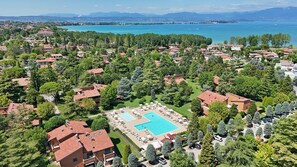 An aerial view of a residential area with a large swimming pool surrounded by lounge chairs and umbrellas, set against a backdrop of a large body of water and mountains. - Holiday Punta Gro in Sirmione (Sirmione)