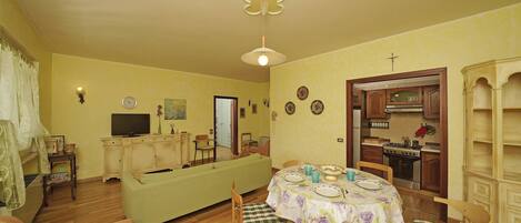 A cozy living room with a sofa, a dining table with chairs, a television, and a kitchen area with cabinets and a stove.