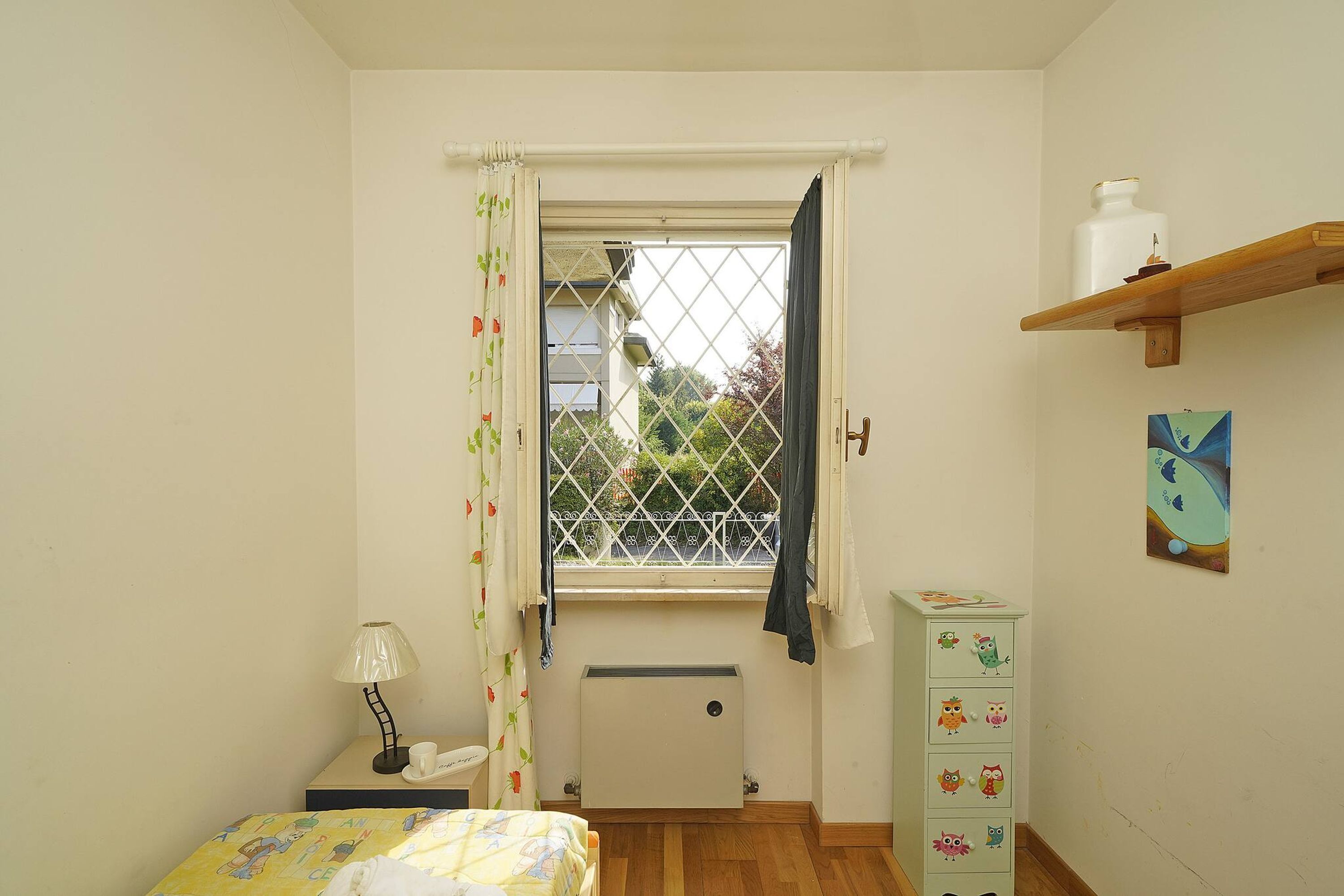 Une petite chambre avec une fenêtre, un radiateur, une table de chevet avec une lampe, un lit recouvert d’une courtepointe colorée, une commode ornée de dessins d’enfants et une étagère avec un vase.