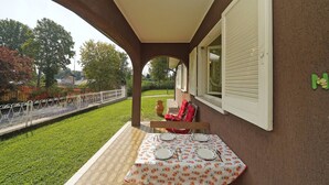 Eine überdachte Terrasse mit Tisch für zwei Personen, einem roten Stuhl und Blick auf Garten und Straße.