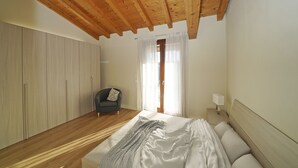 Une chambre avec un plafond en bois, un lit recouvert de linge de lit blanc, un fauteuil gris et une fenêtre ornée de rideaux légers.