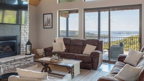 Una sala de estar con chimenea, un sofá de cuero marrón, una mesa de centro y vista a un lago a través de amplios ventanales.