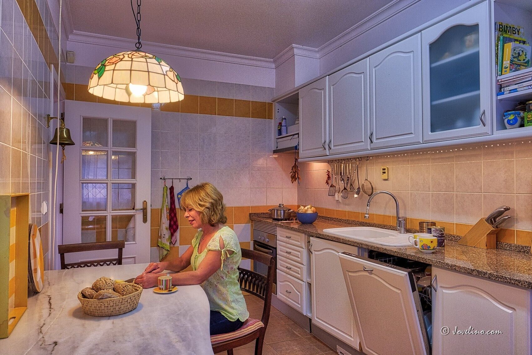 A kitchen with white cabinets, a window, a hanging light fixture, and a dining table with a basket of bread.
