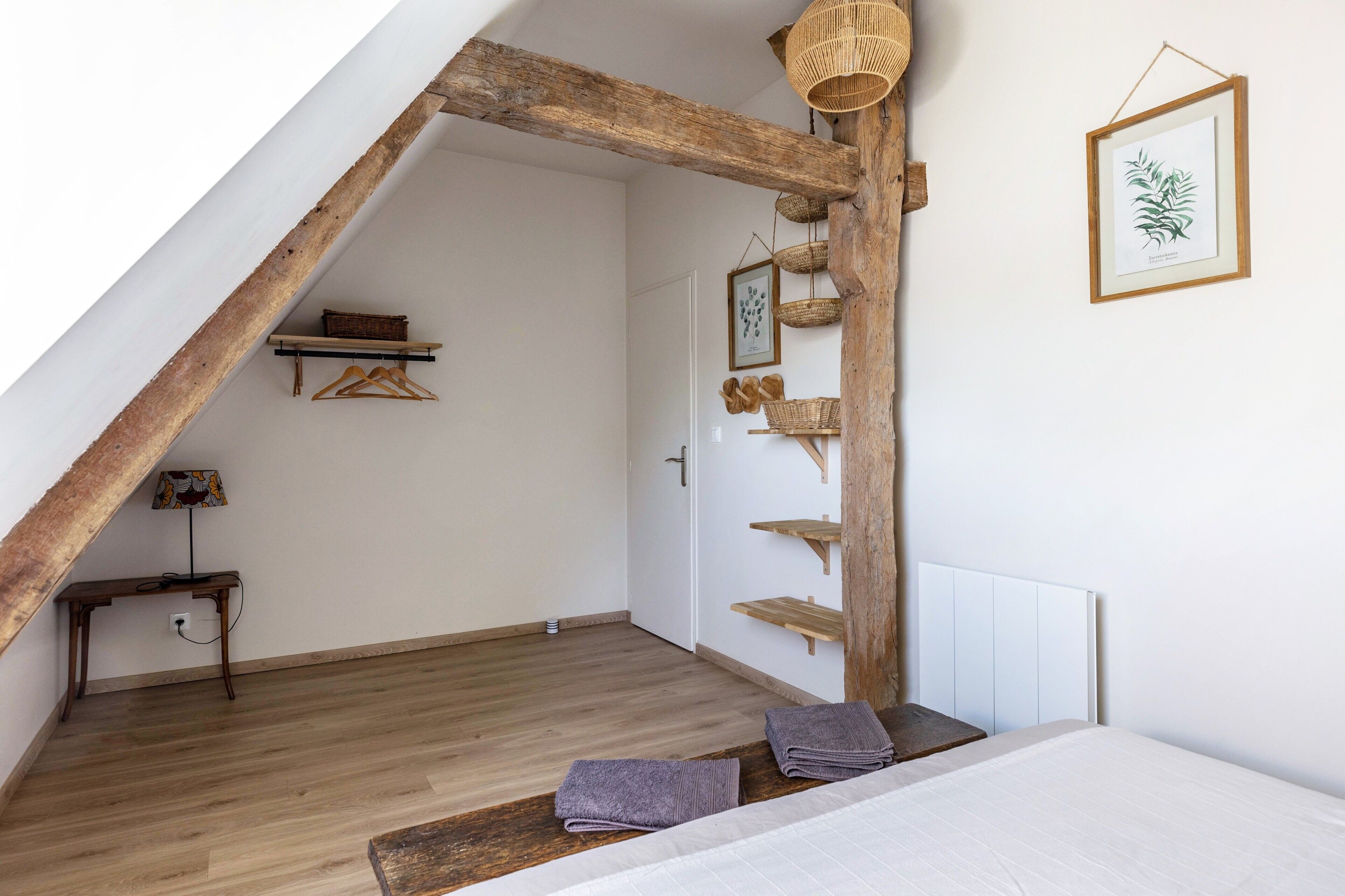 A room with a sloped ceiling, wooden beams, a bed with pillows, a hanging basket, and a framed plant on the wall.