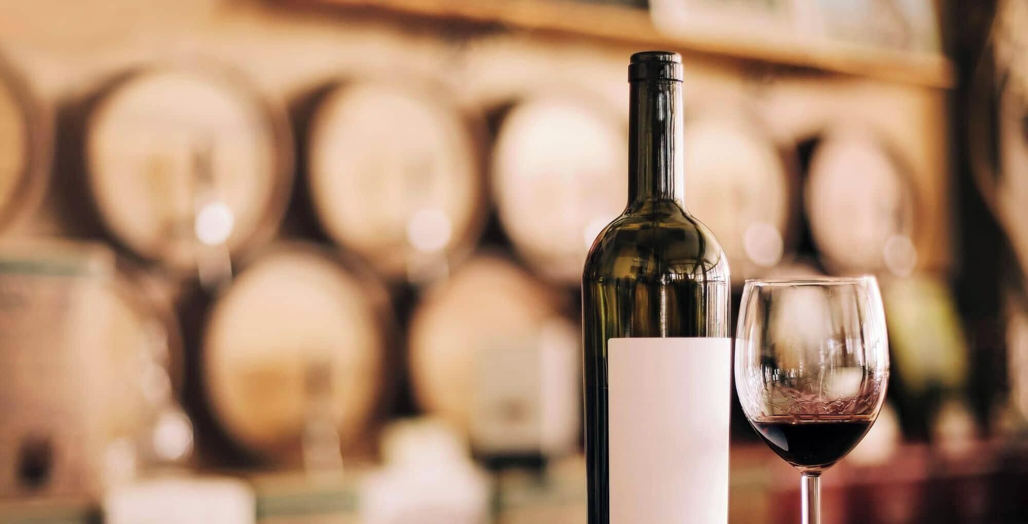 A wine bottle and a wine glass on a counter with a blurred background of wine barrels.