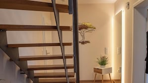 Un intérieur moderne avec un escalier en bois, une petite table blanche agrémentée d’une plante et un luminaire fixé au mur.