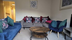 Un salon avec un canapé gris, des fauteuils bleus, une table basse en bois et des coussins à motifs floraux.