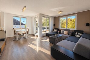 A modern living room with a sectional sofa, a dining area with a table and chairs, and large windows offering views of the outdoors. - Waterkant 20 Apartment (Ostseebad Boltenhagen)