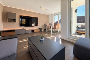 A modern living room with a flat-screen TV, a dining area with a table and chairs, and a large window with a view of buildings. - Waterkant 20 Apartment (Ostseebad Boltenhagen)
