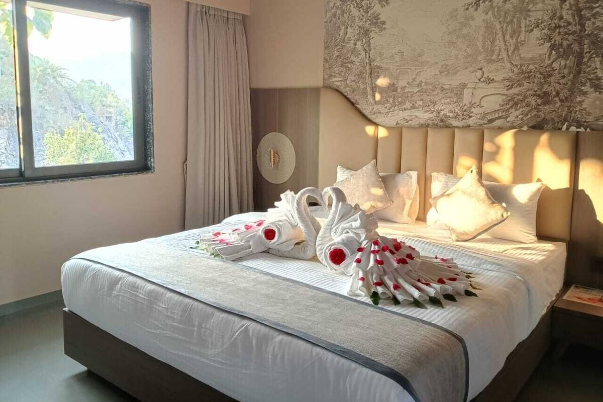 A hotel room with a large bed, a window with curtains, a wall-mounted lamp, and a landscape mural.