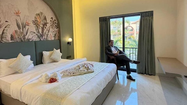 A modern hotel room with a large bed, a ceiling fan, a view of the outdoors, and a painting on the wall.