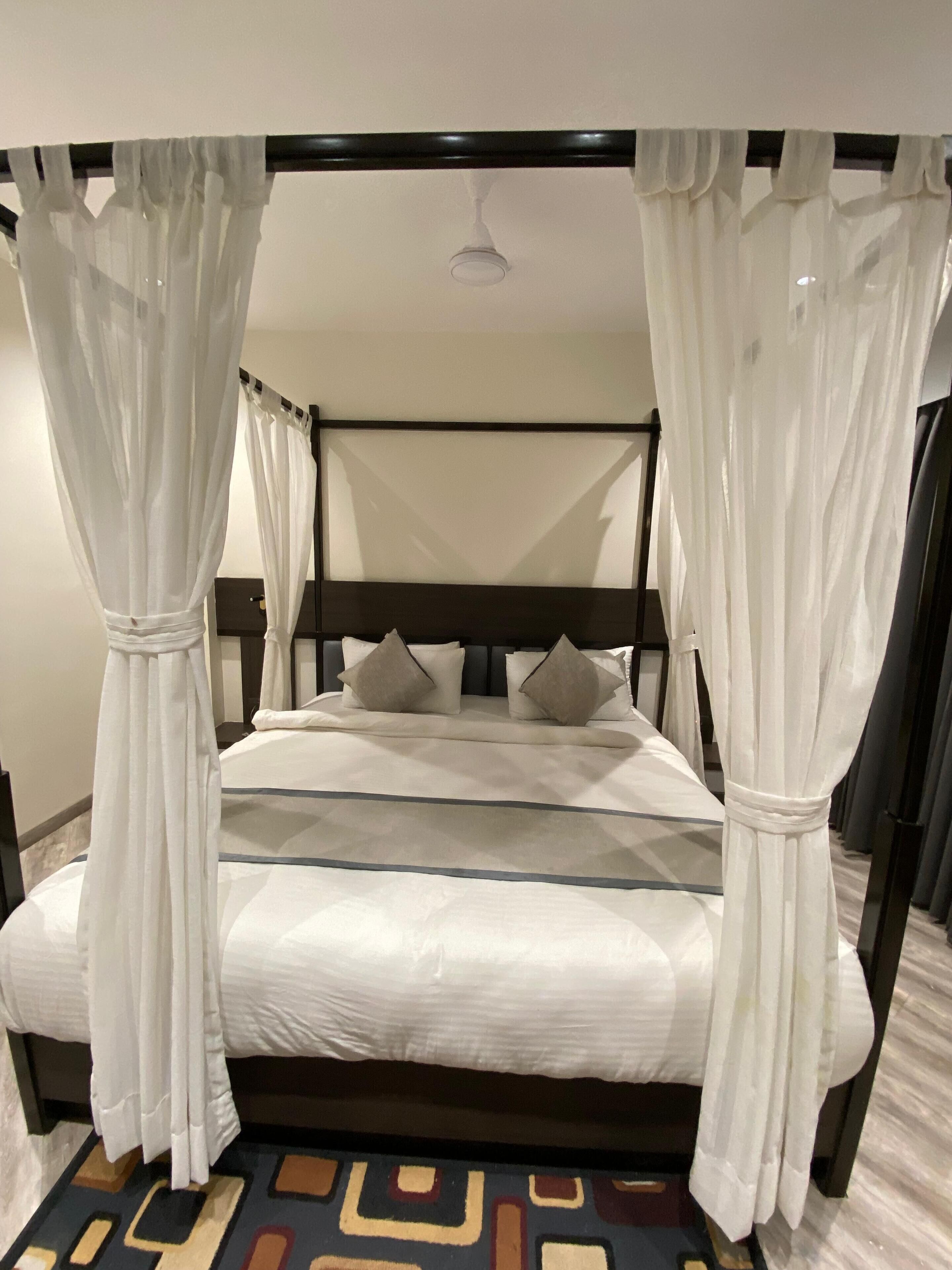A four-poster bed with white bedding and sheer curtains, a dark headboard, and a patterned rug on the floor.