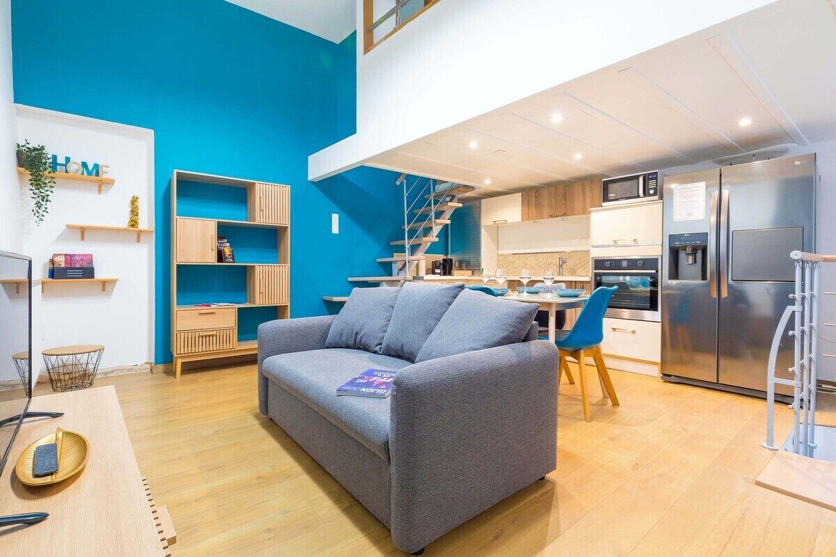 A modern living room with a grey sofa, a dining area with blue chairs, a kitchen with stainless steel appliances, and a blue accent wall.