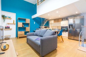 Ein modernes Wohnzimmer mit einem grauen Sofa, einem Essbereich mit blauen Stühlen, einer Küche mit Edelstahlgeräten und einer blauen Akzentwand.
