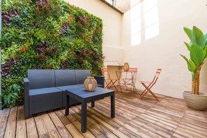 Un patio con un sofá negro, una mesita pequeña y sillas plegables rojas. Se ve una planta grande en maceta a la derecha.