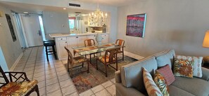 A modern kitchen with white cabinets, a central island, and a chandelier. There is a dining area with a glass table and chairs, and a living room with a sofa and decorative pillows. - Eden E 205 Gulf Front Condo with Stunning Views! (Pensacola)