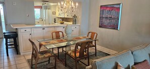 A dining area with a glass table and chairs, a chandelier, and a painting on the wall. - Eden E 205 Gulf Front Condo with Stunning Views! (Pensacola)