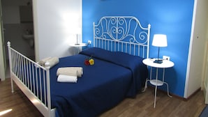 A bedroom with a blue wall, a metal bed frame, a bedside table with a lamp, and a small round table with a chair. - The Blue Room Courtyard (Scicli)