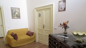 A living room with a yellow sofa, a glass-top coffee table, and a door with a light switch. - The Blue Room Courtyard (Scicli)