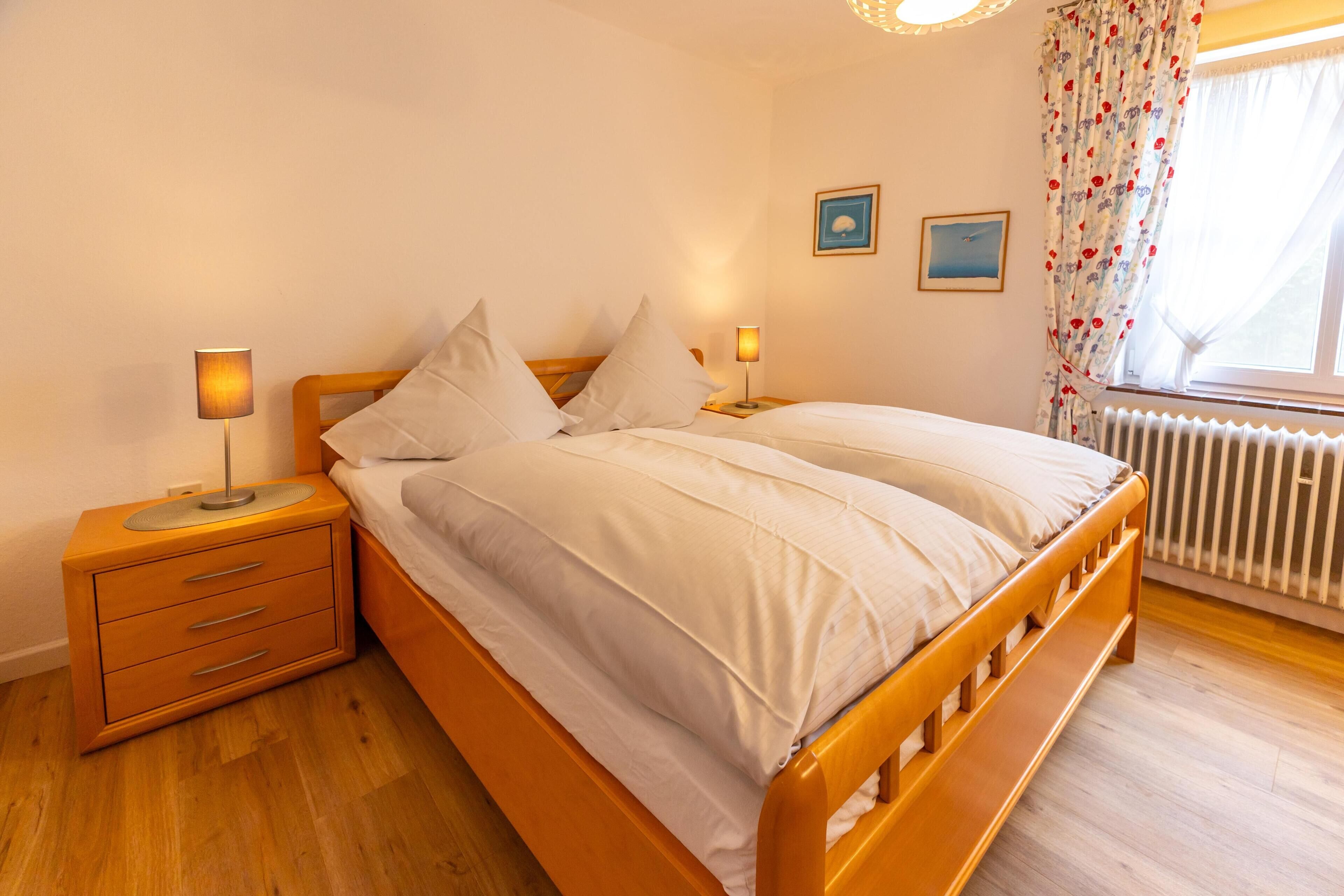 A bedroom with a wooden bed, two bedside tables with lamps, a radiator, and a window with floral curtains.